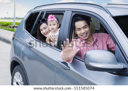 Image of young father waving hands at camera while driving a car with his wife and daughter