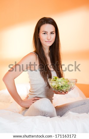 Beautiful young brunette woman eating bowl of salad in bed.