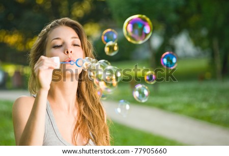 Gorgeous young brunette girl blowing soap bubbles in sunlit park.