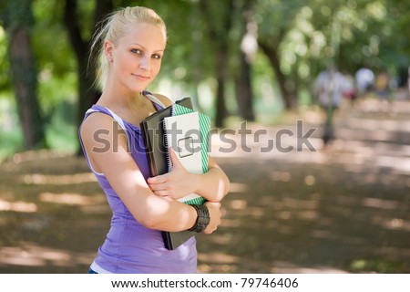 Outdoor portrait of beautiful young blond student girl holding exercise books and laptop.
