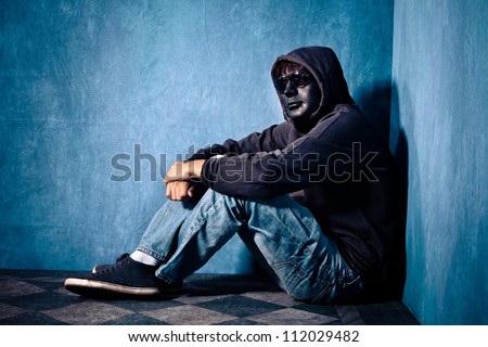 man with black mask sunglasses and hood sit in a corner of blue grunge room full body shot side view