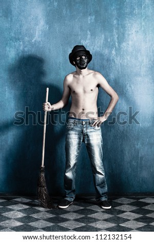 scary man with black mask and hat standing with broom, shirtless in blue jeans in old grunge room