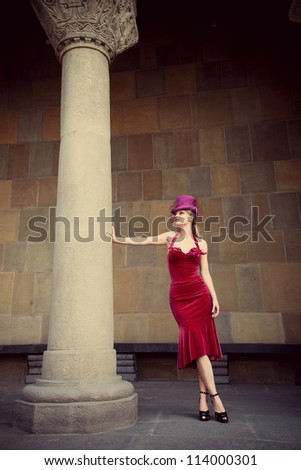 young elegant woman in red dress and cylinder  leaning against column, full body shot