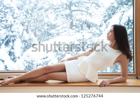 young woman in elegant short dress barefoot sit by window looking at winter snow day