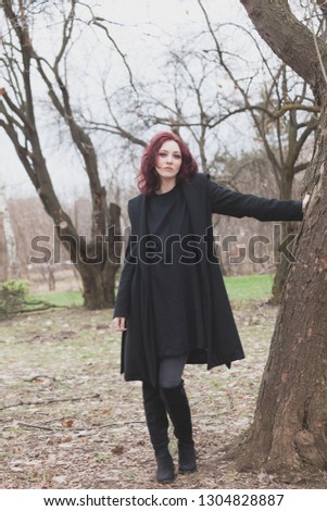 pretty young modern woman in black coat and dress stand by tree in park winter day full body shot