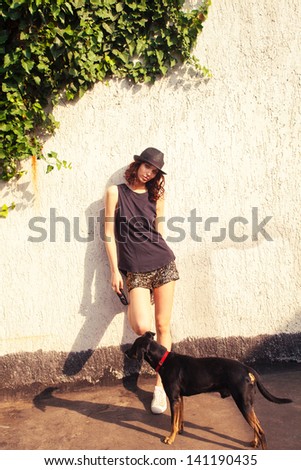 young woman in black top tank, shiny shorts and  hat with dog stand in front wall summer day