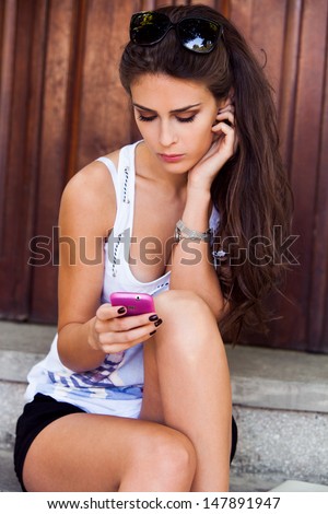 young woman sit on stairs outdoor in casual summer clothes sending message on cell phone