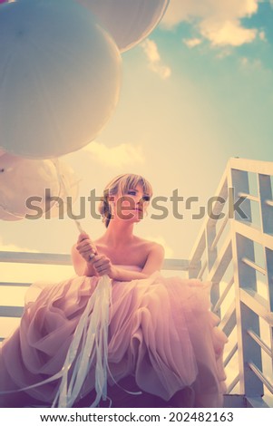 young beautiful woman in elegant wedding dress hold balloons sitting on  white stairs against sky with clouds, retro colors