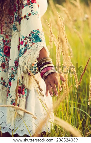 woman wearing boho style clothes touching grass, hand with lot of braceletes, summer day in the field, retro colors