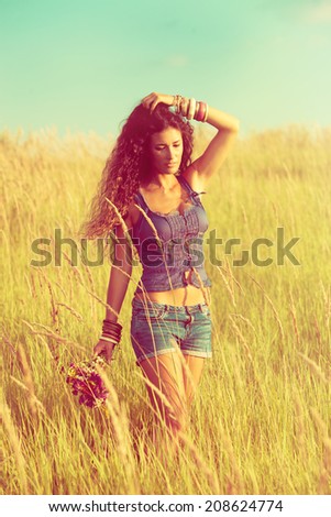 young woman with long curly hair enjoy in walking through yellow summer grass, wearing jeans shorts and boho style top, retro colors