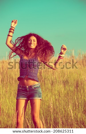smiling young woman with long curly hair jump  in yellow summer grass, wearing jeans shorts and boho style top, retro colors