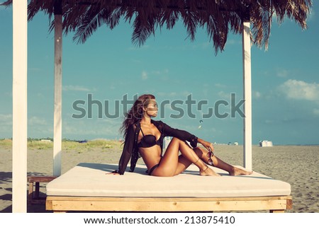 beautiful fashion woman posing on sandy beach in black bikini and shirt sitting on white beach bed under sunshade full body shot