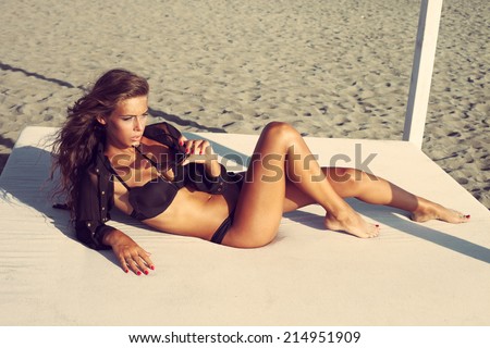beautiful woman posing on sandy beach in black bikini and shirt lie on white beach bed full body shot