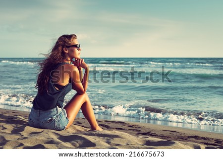 young woman with sunglasses and jeans shorts  sit on sandy beach by the sea enjoy in sunset