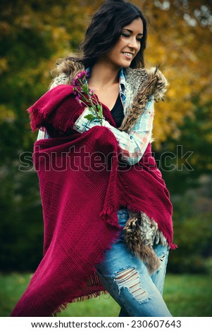 smiling young woman with long curly hair in blue torn jeans, tartan shirt, red wool scarf outdoor shot autumn day in park