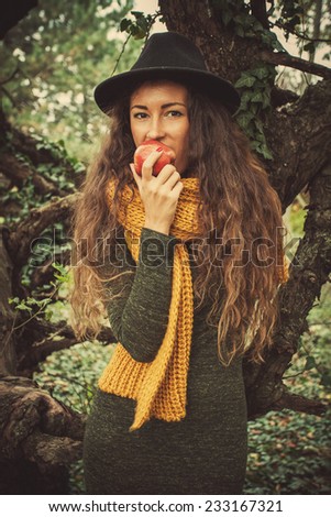 winter autumn girl in wood eat an apple stand by tree wearing hat, wool scarf and green dress