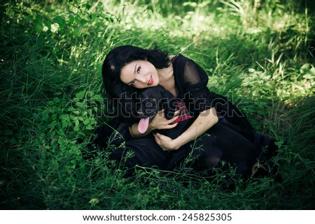 beautiful young woman in black elegant dress sit on grass field with her dog summer day