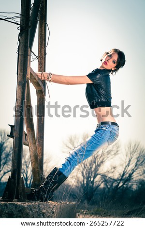 young rebel woman in blue jeans, leather boots and leather jacket outdoor shot on old metal construction, full body shot