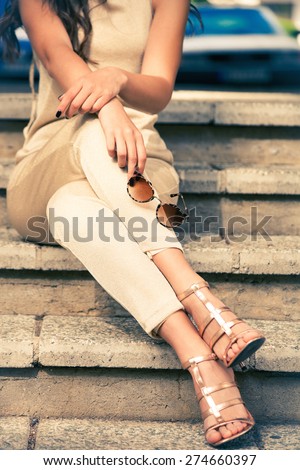 young woman in golden pants and sandals sit on stairs with sunglasses in hand, outdoor shot