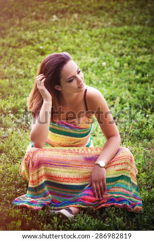 young woman in long colorful dress sit on grass enjoy in warm summer day on meadow