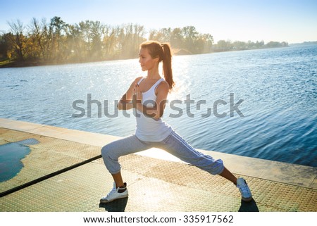 young woman in tracksuit and white undershirt   on pontoon at lake practice yoga, sunny autumn day
