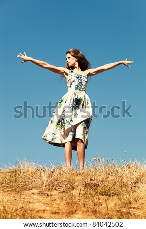 young woman in summer dress with open arms on top of the hill, blue sky in background, summer day