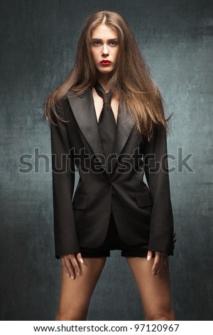 young fashion woman with long brown hair in black tuxedo jacket, tie and shorts, stand in front blue grunge wall