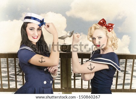 Artistic portrait of two sexy sailor pin up girls with anchor tattoos flexing muscles on a vintage pier promenade when competing in strength and conditioning exercises. Vintage pinup style