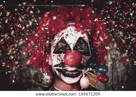 Horizontal Close-Up Portrait On The Face Of A Mad Carnival Clown Holding Balloon Cake Decoration In Mouth Under A Fall Of Confetti. Celebrating Halloween
