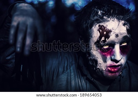 Creepy night photo of a scary zombie looking gravely ill with infectious facial wounds walking through moonlit forest. Attack of the killer monsters