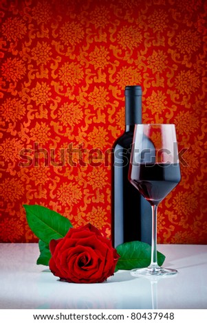 bottle of red wine with half filled glass and red rose on pattern background