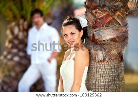 the bride near a palm tree and the groom in the distance. newly married celebrating their honeymoon