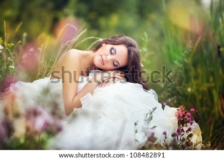 fantastic fairy bride in a wedding dress sits at green grass among flowers with closed eyes.