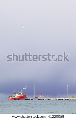 Picture of the pier with a red ship on the background of stormy skies.