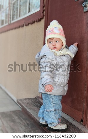 Fun baby knocking at door of house standing on wooden stairs and serious looking back outdoors. Casual baby wanting home after walk