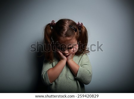 Crying kid girl holding the face the hands and looking down on dark background