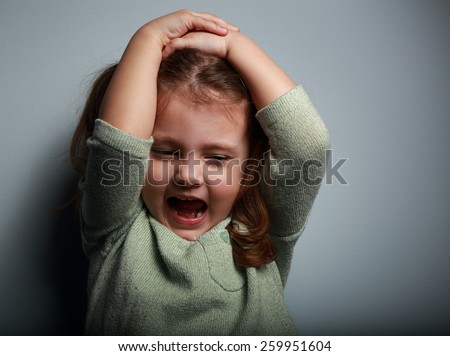 Angry kid girl shouting with open mouth and holding hands the head on dark background