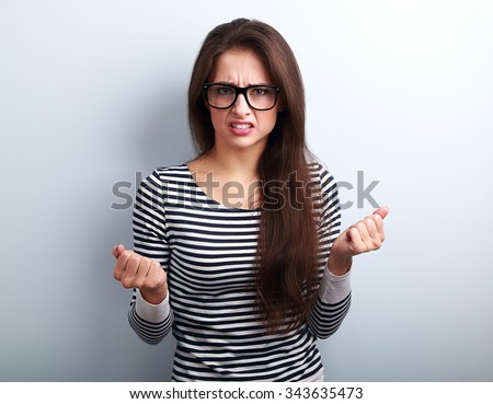 Nervous angry young woman in glasses with aggressive negative face showing fists on blue background