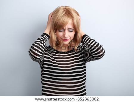 Depressed young blond woman with headache holding head the hands with unhappy face on blue background