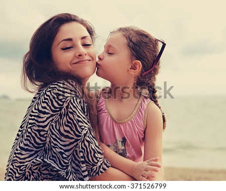 Cute kid girl kissing her happy enjoying mother with closed eyes and emotional face on sea background. Toned color portrait