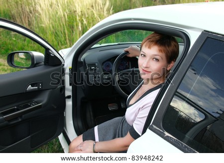 Beautiful smiling woman sitting behind the wheel of sport white car with opened door on nature green background. Sunny day. Good weather