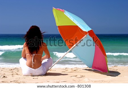 Young, fit, healthy woman sitting under colorful parasol on white sandy beach looking out to sea on a sunny day on vacation.