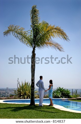 Young couple, in love, holding hands under a palm tree in the sun next to a blue swimming pool in Spain.
