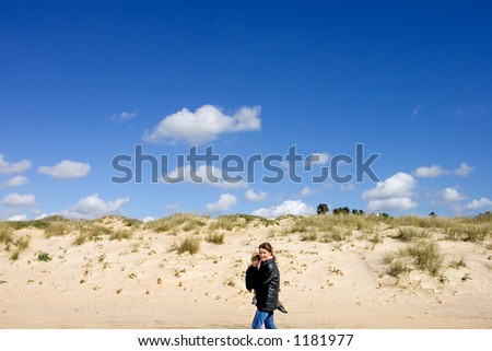 Young mother carrying son and walking along sunny sandy beach in Spain