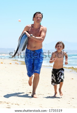 Happy young father and son running along busy, sandy kitesurfing beach with surfboard in the sun