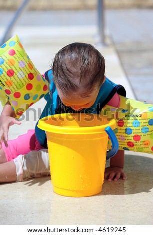 Baby girl sitting next to swimming pool in flotation jacket and arm bands and looking into bucket