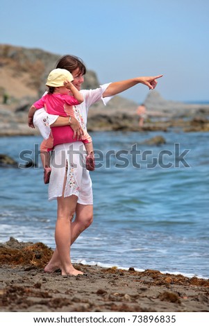 A young mother points to something while carrying her baby daughter on the beach