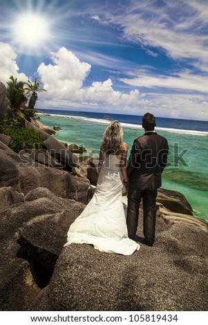Bride and groom standing holding hands on rocks looking out over the ocean at the seashore under tropical sunshine