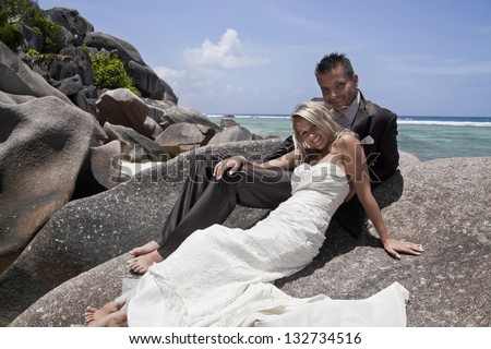Loving beautiful bride and groom posing barefoot in their wedding attire reclining on a rock on the seashore overlooking the ocean