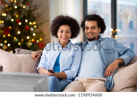 people, family and leisure concept - happy african american couple watching tv at home over christmas tree lights background